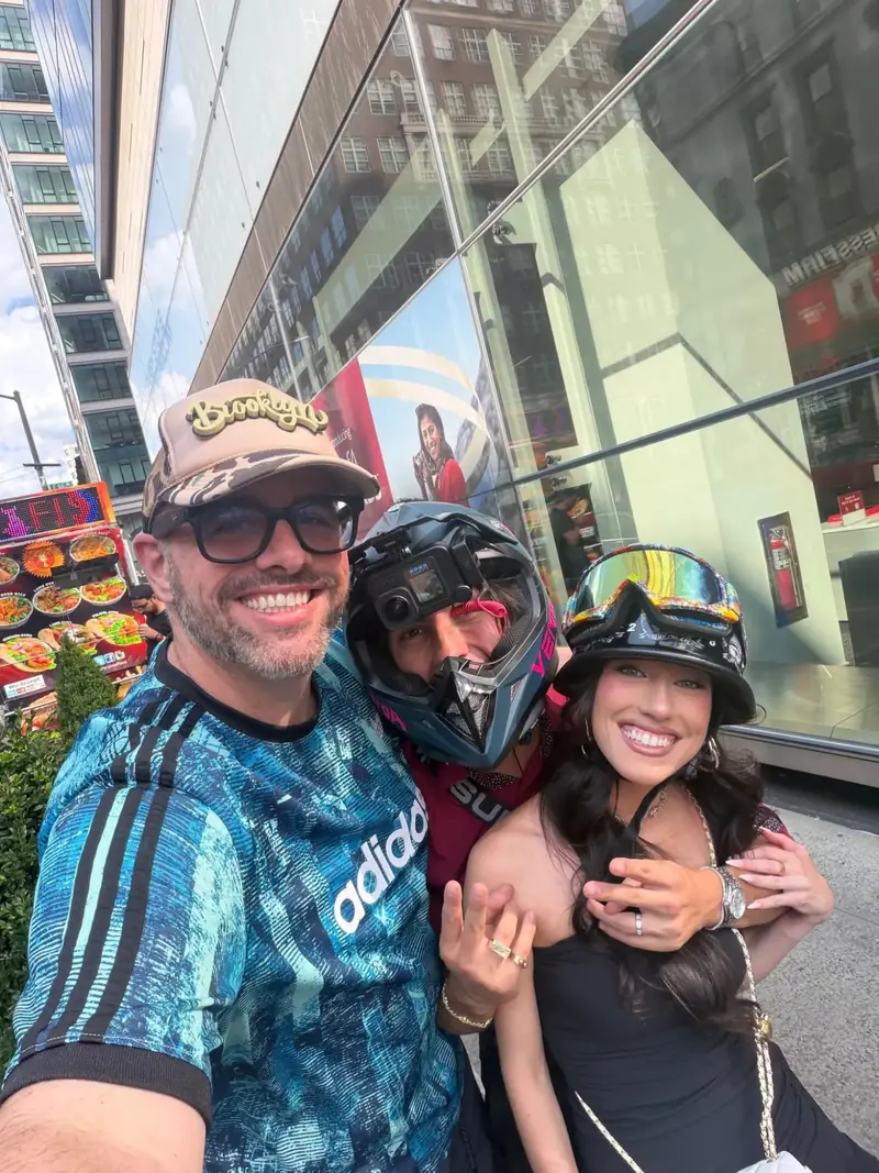 A selfie of three people on a New York street, two of them in motorcycle helmets, in front of a glass building