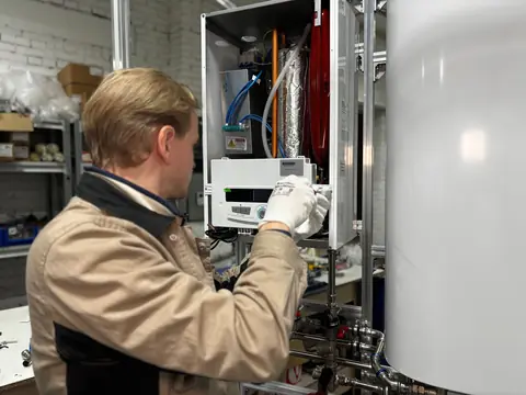 A technician in a tan jacket working inside an open wall boiler, seen from behind