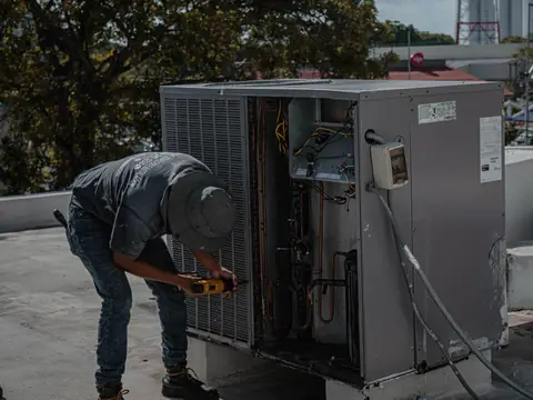 A technician in a sun hat bending over an open air conditioning unit on a flat roof