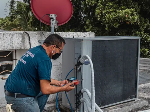 A technician in a face mask crouching with gauges and hoses at an air conditioning unit on a roof