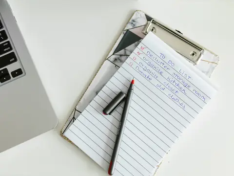 A handwritten to-do list on a clipboard with a pen, beside a laptop