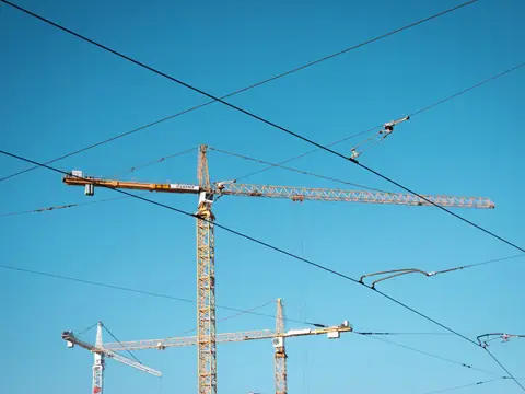 Three tower cranes against a clear blue sky, crossed by overhead wires