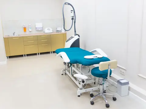 A white treatment room with a teal treatment bed, a stool and a machine beside gold cabinets
