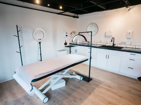 A treatment room with a white treatment table, a ring light and white cabinets on a wood floor