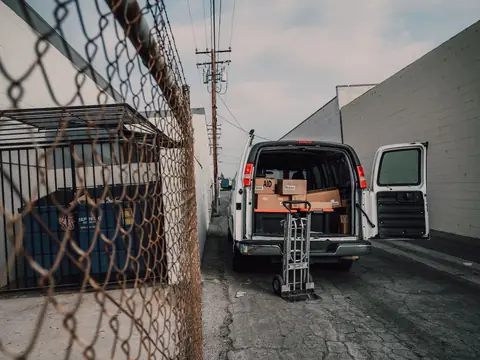 A white van with its back doors open and boxes inside, a hand truck behind it, seen along a wire fence