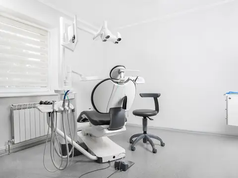 A white dental room with a treatment chair, a stool and a lamp by a window