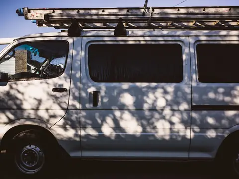 A silver work van with ladders on its roof rack, parked in the shade of a tree