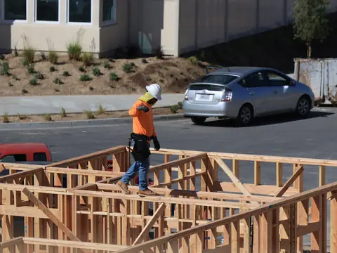 A worker in an orange vest and hard hat walking on the timber framing of a house