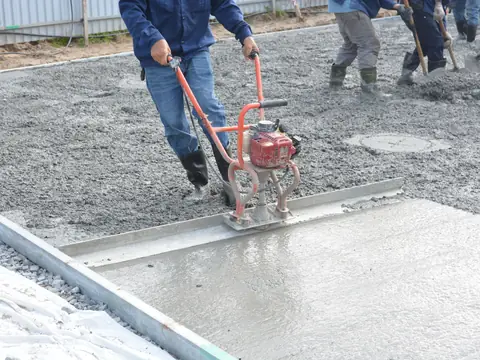 A worker drawing a powered screed across wet concrete while others shovel behind him