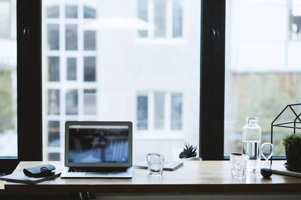 A modern office desk with a laptop, a glass of water, and a view through an open window.